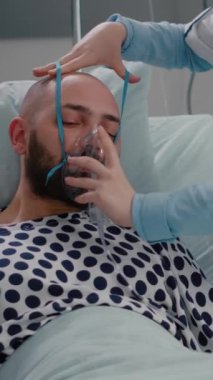 Dikey video: Kadın doktor oksijen maskesi takıp solunum hastalığını izlerken hasta yatakta oturuyor. Doktor, iyileşme sürecinde hastane koğuşunda hastalık tedavisi görüyor.