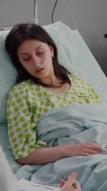 Dikey video: Doktor pratisyen hekimin hastane koğuşundaki iyileşme randevusu sırasında nabzını kontrol etmek için panoya hastalık belirtileri yazması. Hasta kadın hasta, yatakta dinleniyor.
