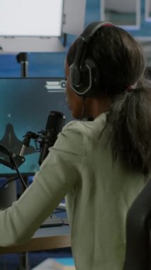 Dikey video: Siyah oyuncu kulaklık takıyor diğer oyuncularla sohbet ediyor ve online turnuvada profesyonel nişancı oyunları oynuyor. Oyuncu yeni grafiklerle çevrimiçi video oyunları yapıyor