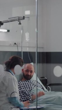 Dikey video: Hastanın hayati organlarını kontrol eden tıbbi asistan kalp atışlarını takip ediyor hastane koğuşunda serum serumuna vitamin enjekte ediyor. Doktor, tıbbi tedaviyi tartışıyor.
