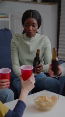 Dikey video: Çok kültürlü arkadaşlar kanepede dinlenirken birlikte zaman geçirdikleri bira şişelerini alkışlıyorlar. Bir grup melez insan gece geç saatlerde wekeend partisinde takılıyorlar, sosyalleşiyorlar.