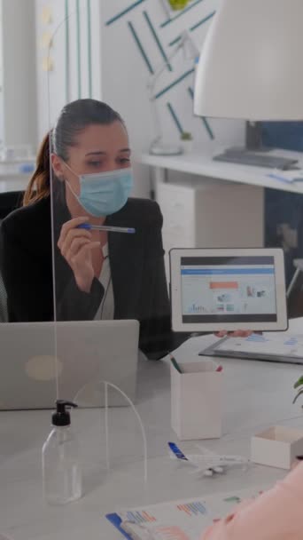 垂直ビデオフェイスマスクを持つビジネスマンは デジタルタブレットコンピュータを使用して彼女の同僚との統計を分析します 社会的距離を尊重する新しい通常のオフィスで働くチームは感染を防ぐために — ストック動画