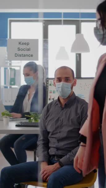 垂直ビデオコロナウイルスのパンデミックに対する保護面マスクを身に着けている起業家の男の肖像 オフィスのテーブルデスクで働く従業員は ウイルスを防ぐために社会的距離を維持します — ストック動画
