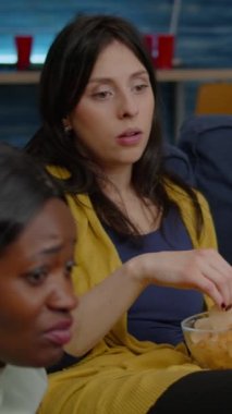 Dikey video: Çok ırklı arkadaşlar dizüstü bilgisayarda online film izliyorlar kanepede dinleniyorlar ve birlikte gülüyorlar. Bir grup çok ırklı insan gece geç saatlerde takılıyor, bira içiyor, abur cubur yiyor.