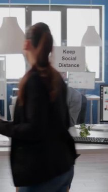 Koruyucu yüz maskesi takan dikey video Freelancer tıbbi termometre kullanarak takım sıcaklığını kontrol ediyor. İş arkadaşları enfeksiyondan kaçınmak için yeni normal şirket ofisinde sosyal mesafeye saygı gösteriyorlar.