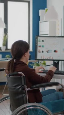Dikey video: Şirketin yıllık istatistiklerini kontrol etmek için son teslim tarihi için iş ofisinde çalışan etkisiz hale getirilmiş engelli yönetici kadın. Modern ağı kullanarak tekerlekli sandalyedeki çalışana odaklan