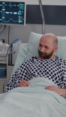 Dikey video: Hasta adam hasta yatağında dinleniyor oksijen tüpü takarak pratisyen doktora hastalık belirtilerini açıklarken panoya hastalık tedavisi yazıyor. Tıp hemşiresi oksimetreyi inceliyor.