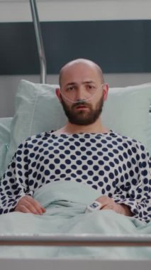 Dikey video: Hastane koğuşunda iyileşme sürecinde nazal oksijen tüpü takmış, hasta bir adamın kameraya kötü baktığı potrait. Solunum tedavisi bekleyen depresif bir hasta.