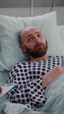 Dikey video: Kadın hemşire kalp atışlarını kontrol ederken, siyah derili bir doktor panoya tıbbi uzmanlık yazısı yazıyor. Hasta nazal oksijen tüpüyle kötü bir şekilde dinleniyor.