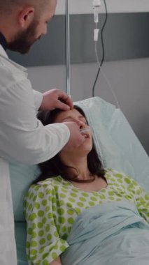 Dikey video: Hastane koğuşunda koronavirüs salgını sırasında oksijen maskesi takan hasta bir kadını izleyen klinik ekip. Siyah hemşire kalp atışlarını kontrol ediyor.