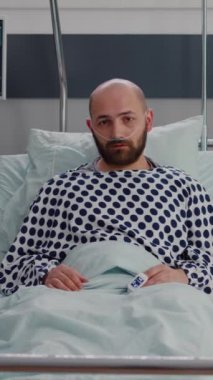Dikey video: Hastane koğuşunda ameliyat sonrası iyileşme sürecinde olan hasta bir adamın yatakta dinlenip solunum tedavisini beklemesi. Hastanedeki hasta nazal oksijen kullanan kameraya bakıyor.