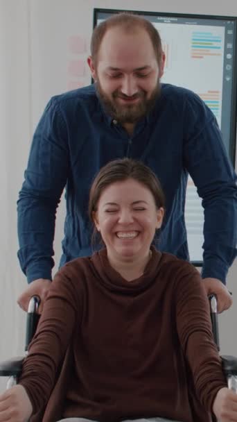 视频垂直 快乐的员工带着瘫痪的残疾同事笑着坐在轮椅上休息 兴致勃勃地创办了财务办公室 不动声色的女人笑 — 图库视频影像