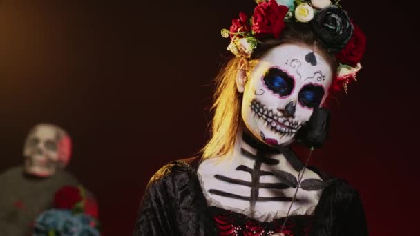 死の衣装と花を身に着けている美しいモデルは スタジオの背景に花輪を捧げ 聖なるサンタ マエルテのように振る舞っています 頭蓋骨のボディアートで死んだラ カヴァレラ カトリーナの女性 メキシコ文化 手持ち撮影 — ストック動画