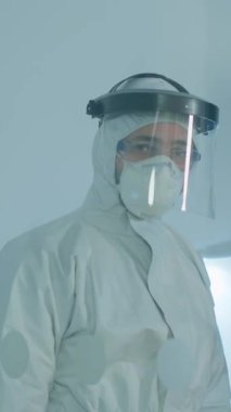 Dikey video: Coperid-19 'a karşı koruyucu giysi giyen yorgun doktor tehlike bölgesinde çalışan cam duvarın arkasındaki kamerada bitkin görünüyor. Bilim adamı yüksek teknoloji kullanarak virüs evrimini inceliyor.