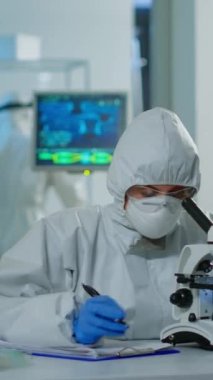 Dikey video: Tıbbi araştırma bilim adamı mikroskop altında DNA deneyleri yapıyor koruyucu giysi giyiyor ve panoya bilgi yazıyor. Kimyager aşıyı geliştirmek için yüksek teknoloji kullanıyor.