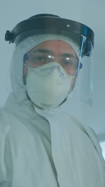 Dikey video: Teçhizatlı laboratuvar elbisesi içinde mikrobiyolog laboratuvarda durup cam duvarın arkasındaki kameraya bakıyor. Yüksek teknoloji ve kimya araçları kullanarak virüs evrimini inceleyen doktor