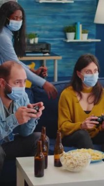 Dikey video: Coronavirus yayılmasını önlemek için oturma odasında joystick kontrolörüyle video oyunu oynayan konsantre olmuş çok ırklı arkadaşlar. Çeşitli insanlar eğleniyor, gülüyor.