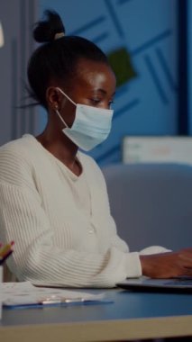 Projenin son teslim tarihine uymak için yeni normal iş ofisinde dizüstü bilgisayarda fazla mesai yapan koruma maskeli Afrikalı bir çalışan, küresel salgın sırasında masa başında fazla mesai yapan belgeleri analiz ediyor.