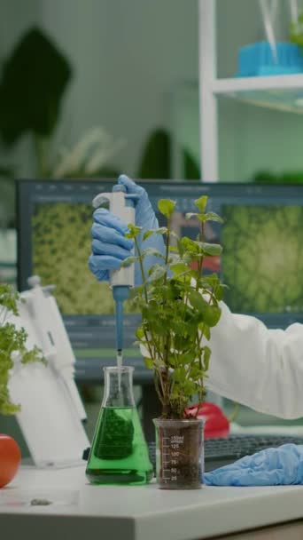 垂直ビデオ 遺伝学実験のための緑の苗木を置く医療用フラスコからの生物学者の話の解決策 専門の微生物学研究所で働く白衣の女性研究者 — ストック動画