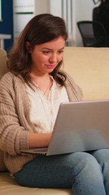 Dizüstü bilgisayarda rahatlama bölgesinde oturan, rahat koltukta oturan, bilgisayar başında gülümseyen ve arka planda çalışan çeşitli meslektaşları olan kadın yönetici. Yeni finans projesi planlayan çok ırklı iş arkadaşları