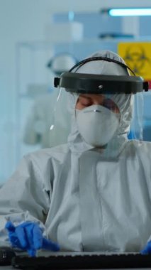 Dikey video: ppe suit 'teki laboratuvar teknisyeni pc' deki test tüpündeki kan örneğini analiz ediyor. Doktor çeşitli bakteriler, doku ve antibiyotikler için ilaç araştırmaları yapıyor.