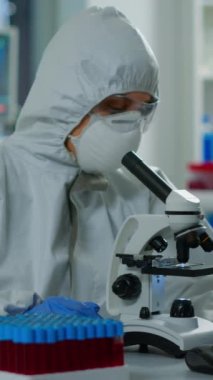 Dikey video: Kıyafetli kadın bilim adamı labaratuarda slaytlarla modern mikroskop kullanarak çalışıyor. Kimyagerlerden oluşan bir ekip virüs evrimini araştırıyor. Bilimsel araştırmalar için yüksek teknoloji kullanıyor.