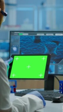 Dikey video: Modern donanımlı laboratuvarda, krom anahtar ekranlı tableti tutan ve inceleyen araştırmacı. Aşı araştırması yapan mikrobiyologlardan oluşan bir ekip yeşil ekranlı, izole edilmiş bir cihaz üzerinde yazıyor.
