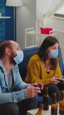 Dikey video: Üzgün arkadaşlar çünkü sosyal salgın zamanında Corona 'ya yakalanmamak için kablosuz kontrolörle oyun oynamak yüz maskesi takmak. Bir grup insan eğleniyor.