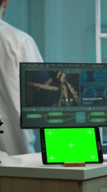 Dikey video: LaBooratuarda çalışan yeşil ekranlı tablet, arka planda profesyonel mühendis virüs evrimini test ederken krom anahtar görüntüsü. Kanlı yüksek teknoloji ürünü bir laboratuvar.