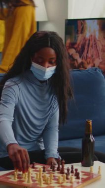 Dikey video: Çeşitli arkadaşlar, küresel salgın sırasında yüz maskesi takan Covid19 hastalığını önlemek için evde satranç oynayan bira şişelerini tokuşturarak sosyal mesafeyi koruyorlar. Karışık insanlar eğleniyor