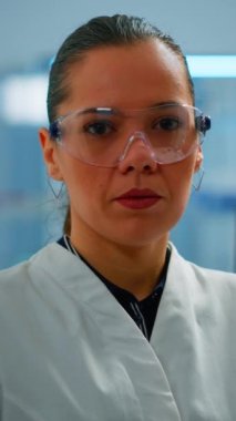 Dikey video: Modern donanımlı laboratuvarda iç çeken kameraya bakan yorgun kimyager doktor. Bilimsel araştırma ve aşı için yüksek teknoloji ve kimya araçları kullanarak virüs evrimini inceleyen çok ırklı bir ekip.
