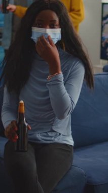 Dikey video: Afrikalı kadın yüz maskesi takarak sosyal mesafeyi koruyor Coronavirus 'un yayılmasını önlemek için arkadaşlarıyla buluşuyor elinde bira şişesi, koltukta oturan kameraya bakıyor, covid 19