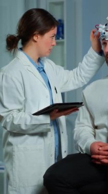 Dikey video: Beyin dalgası tarama kulaklığıyla nörolojik sandalyede oturan adam, hasta beyninin evrimini tablet kullanarak takip eden araştırmacı doktor, eeg cihazını ayarlıyor. İnceleme yapan bilim adamı