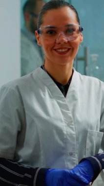 Dikey video: Laboratuvar doktoru modern donanımlı laboratuvarda gülümseyen kameraya bakıyor. Bilimsel araştırma ve aşı için yüksek teknoloji ve kimya araçları kullanarak virüs evrimini inceleyen çok ırklı bir ekip.