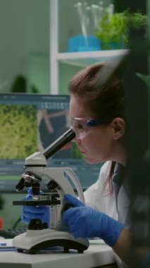 Dikey video: İlaç bilimcisi not defterine genetik test yazarken mikroskopta yeşil yaprak örneğine bakıyor. Biyolog, çalışırken organik gmo bitkilerini analiz ediyor.