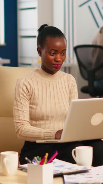 バックグラウンドで多様なチームが働いている間 カメラの笑顔を見てラップトップに入力するアフリカの女性の肖像画 現代のビジネスオフィスでスタートアップ金融会社について話す多民族の同僚 — ストック動画