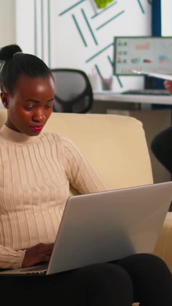 专心致志的非洲商界女性在繁忙的创业办公室里坐在沙发上回复在笔记本电脑上打字的电子邮件 而多样化的团队则分析统计数据 在新项目中工作的多族裔小组 — 图库视频影像