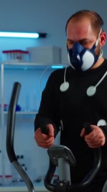 Dikey video: Maskeli ve elektrotlu sporcu bilim sporları laboratuarının sağlık durumunu izleyen çapraz eğitimciye bağlı. Performans kardiyosunu ölçen tıbbi araştırmacı.