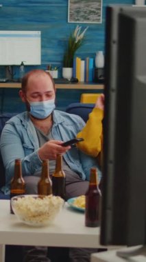 Dikey video: Küresel salgın sırasında koltukta oturup eğlenirken, küresel salgın sırasında yüz maskesi takarak gülen bir grup çok ırklı arkadaş televizyonda komedi programı izliyor.