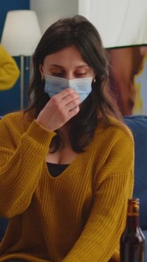 Dikey video: Koruma maskeli genç bayan oturma odasında kanepede oturmuş kameranın önünde bira içiyor. İnsanlar covid 19 salgını sırasında sosyalleşiyorlar sosyal mesafeye saygı gösteriyorlar, eğleniyorlar.