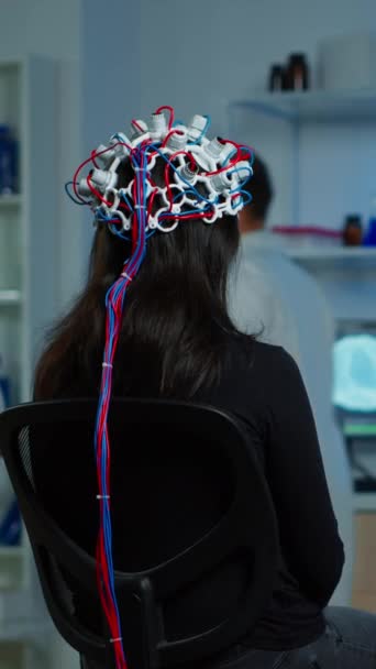 垂直ビデオ 医療研究者がそれを調整しながら 神経研究室で椅子に座ってパフォーマンスの目のヘッドセットを身に着けている女性患者のバックビュー 上の神経系のタイプを調べます — ストック動画