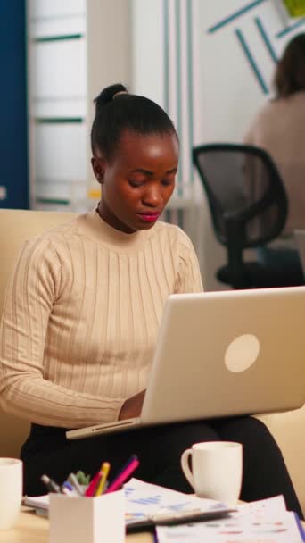 アフリカ系アメリカ人の女性がカメラを見てノートパソコンに書き込み 多様な同僚が背景で働いている 現代のビジネスにおけるスタートアップ金融会社について話す多民族の同僚 — ストック動画