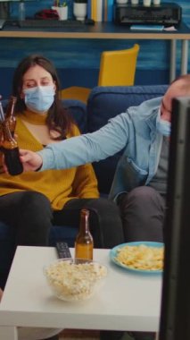 Dikey video: Karışık ırklar televizyon izliyor, sosyalleşiyor, bira şişelerine tezahürat yapıyor, birlikte vakit geçiriyorlar sosyal mesafeye saygı gösteriyorlar ve covid 19 virüsüne karşı koruyucu maskeler takıyorlar. Rahatlatıcı...