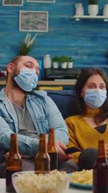 Dikey video: Çeşitli insanların birlikte oturup uzaktan kumandalı televizyon izlemesi, Coronavirus salgını sırasında maske takarak hastalıkları önlemesi. Partinin tadını çıkarıyor bira içiyor.