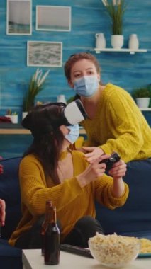 Dikey video: Video oyunları oynayan kadın VR kulaklık takıyor ve sosyal salgın sırasında sosyal mesafeyi korumasına rehberlik eden çok ırklı arkadaşlar. Çeşitli insanlar yeni normal bir partide eğleniyor.