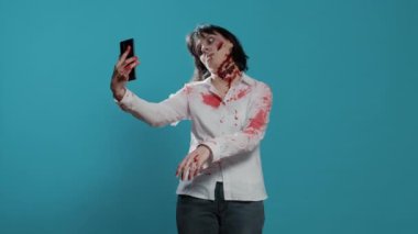 Yürüyen bir ölü, mavi arka planda akıllı telefon cihazıyla selfie çekiyor. Ürkütücü ve korkunç görünümlü kadın. Derin ve kanlı yaraları olan ve kendi fotoğrafını çeken..