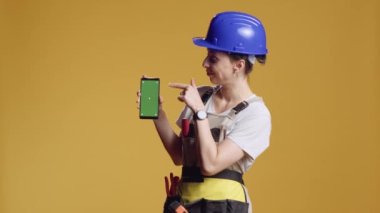 Güçlü kadın müteahhit, sarı arka planda duran akıllı telefondan yeşil ekran ekranını işaret ediyor. Model şablonu ve krom anahtarı ile izole edilmiş telif alanı kullanmak, sanayi inşa etmek.