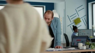 Man in startup office explaining sales charts and statistics to woman coworker taking notes standing over desk. Colleagues talking about company turnover over table with business papers and laptop.