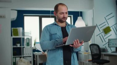 Portrait of casual business man holding laptop typing on keyboard and smiling at camera in busy modern office. Confident startup employee working relaxed in finance department of small company.