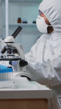 Dikey video: Ekipmanlı laboratuarda mikroskopla inceleyen bilim adamının kostüm giymesi. Ekip, aşı geliştirme araştırmalarında yüksek teknoloji kullanarak virüs evrimini inceliyor.